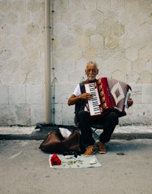 An elderly man is sitting on a chair on the street playing a red accordion. He is positioned against a textured stone wall. There is a bag and some coins placed on a cloth on the ground beside him.