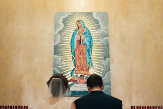 A couple is seen from behind, standing in front of a religious mosaic artwork depicting the Virgin Mary. The Virgin is shown in traditional attire with a blue mantle and red dress, surrounded by radiant beams and clouds. The setting appears to be a solemn and sacred space.
