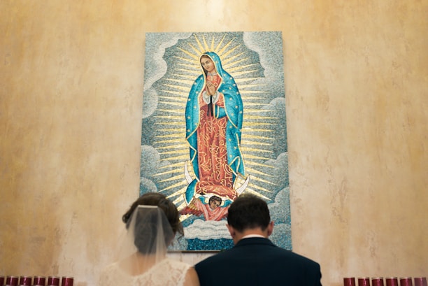 A couple is seen from behind, standing in front of a religious mosaic artwork depicting the Virgin Mary. The Virgin is shown in traditional attire with a blue mantle and red dress, surrounded by radiant beams and clouds. The setting appears to be a solemn and sacred space.