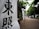 Vertical banners with bold black Japanese characters are displayed along a path bordered by lush green trees. The scene suggests a tranquil, outdoor setting, possibly leading to a significant location or event.