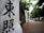 Vertical banners with bold black Japanese characters are displayed along a path bordered by lush green trees. The scene suggests a tranquil, outdoor setting, possibly leading to a significant location or event.