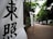 Vertical banners with bold black Japanese characters are displayed along a path bordered by lush green trees. The scene suggests a tranquil, outdoor setting, possibly leading to a significant location or event.