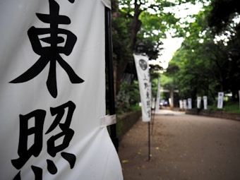 Vertical banners with bold black Japanese characters are displayed along a path bordered by lush green trees. The scene suggests a tranquil, outdoor setting, possibly leading to a significant location or event.