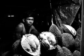 A man sits in a dimly lit space surrounded by a pile of durians. The light highlights the texture of the fruit's spiky skin and casts shadows on the man's face. The setting appears rustic and intimate.