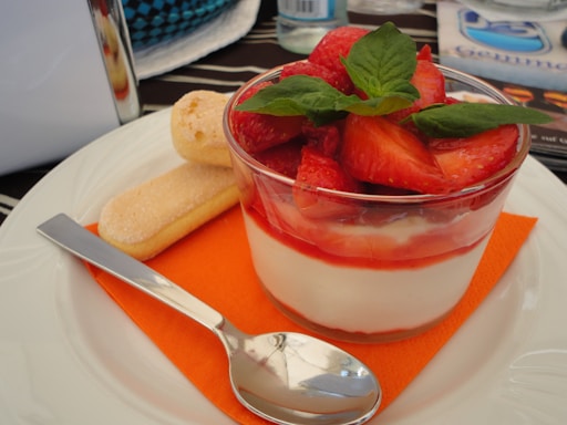 A dessert consisting of a glass bowl filled with creamy white yogurt or mousse, topped with fresh strawberries and mint leaves. It is placed on an orange napkin with a silver spoon and ladyfinger biscuits beside it, all set on a white plate. The background includes patterned objects and a water bottle.