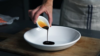 person dripping black liquid from small white ceramic bowl to big white ceramic bowl sauce teams background