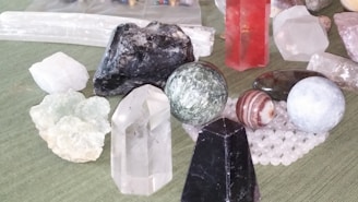 A collection of various gemstones and crystals placed on a surface. The assortment includes different shapes such as spheres, prisms, and raw stones. Notable colors include clear, black, red, and green. Some of the stones are polished, while others remain in their raw form.