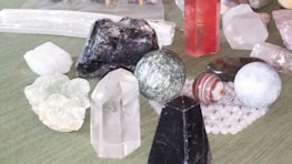 A collection of various gemstones and crystals placed on a surface. The assortment includes different shapes such as spheres, prisms, and raw stones. Notable colors include clear, black, red, and green. Some of the stones are polished, while others remain in their raw form.