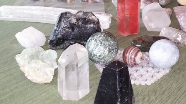 A collection of various gemstones and crystals placed on a surface. The assortment includes different shapes such as spheres, prisms, and raw stones. Notable colors include clear, black, red, and green. Some of the stones are polished, while others remain in their raw form.