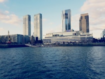 A waterfront scene with modern high-rise buildings in the background and a river in the foreground. The architecture is contemporary, featuring a hotel with the Hilton logo visible. The sky is partly cloudy, adding to the urban cityscape vibe.