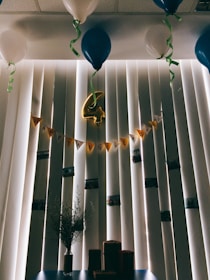 A room decorated for an anniversary celebration, featuring a string of yellow pennant flags spelling 'Happy Anniversary' against vertical blinds. Blue and white balloons are tethered to the ceiling, with green ribbons hanging down. In the center, a gold foil balloon in the shape of the number four is hanging. Below, a vase with sprigs of greenery sits on a table alongside several small wrapped packages.