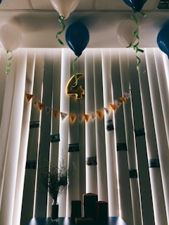 A room decorated for an anniversary celebration, featuring a string of yellow pennant flags spelling 'Happy Anniversary' against vertical blinds. Blue and white balloons are tethered to the ceiling, with green ribbons hanging down. In the center, a gold foil balloon in the shape of the number four is hanging. Below, a vase with sprigs of greenery sits on a table alongside several small wrapped packages.