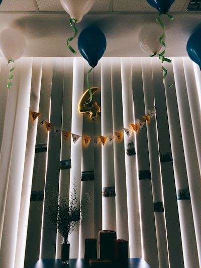 A room decorated for an anniversary celebration, featuring a string of yellow pennant flags spelling 'Happy Anniversary' against vertical blinds. Blue and white balloons are tethered to the ceiling, with green ribbons hanging down. In the center, a gold foil balloon in the shape of the number four is hanging. Below, a vase with sprigs of greenery sits on a table alongside several small wrapped packages.