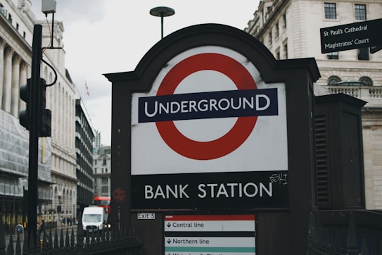 An entrance to a London Underground station marked by the iconic red and blue roundel is displayed prominently, surrounded by classical architecture of the city. Nearby, street signs point to notable locations such as St Paul's Cathedral and the Magistrates' Court. A red double-decker bus is visible in the background, emphasizing the urban setting.