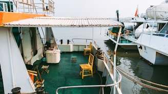 A boat with a covered deck is moored next to another vessel. The deck is equipped with several yellow chairs and a small table. There are ropes tying the boat to a dock and various nautical equipment visible. The setting appears to be a harbor or marina.