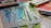 A collection of crafting materials including colorful sequins, rectangular metal frames with embossed patterns, alcohol markers, colored pencils in shades of blue, and a black ink pad. A piece of paper with watercolor artwork is partially visible alongside a whimsical cartoon sketch.