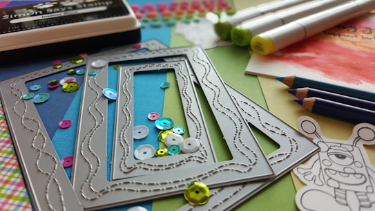 A collection of crafting materials including colorful sequins, rectangular metal frames with embossed patterns, alcohol markers, colored pencils in shades of blue, and a black ink pad. A piece of paper with watercolor artwork is partially visible alongside a whimsical cartoon sketch.