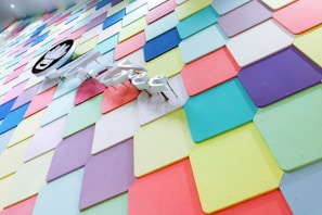 A vibrant wall made of colorful, overlapping rectangular panels arranged in a diamond pattern. The panels display a variety of pastel shades including blue, pink, yellow, and purple. A circular sign with a monochrome design is mounted on the wall, alongside a neon sign that appears to resemble text.
