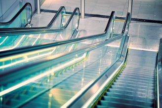 A modern escalator with a sleek design extends upwards, featuring transparent glass sides and illuminated yellow edges. The setting appears to be a contemporary indoor space with a polished surface surrounding the structure.