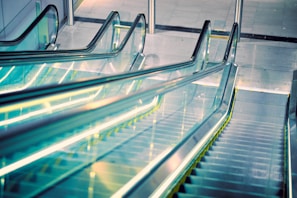 A modern escalator with a sleek design extends upwards, featuring transparent glass sides and illuminated yellow edges. The setting appears to be a contemporary indoor space with a polished surface surrounding the structure.