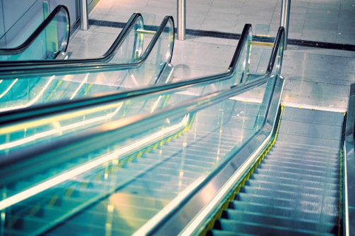 A modern escalator with a sleek design extends upwards, featuring transparent glass sides and illuminated yellow edges. The setting appears to be a contemporary indoor space with a polished surface surrounding the structure.