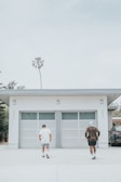 Two people are seen walking towards a modern garage with two large doors. The garage is part of a white building with minimalistic design features. There's a lone palm tree in the background and a parked vehicle partially visible to the right. The atmosphere appears calm, suggesting a casual and relaxed setting.