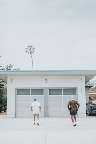 Two people are seen walking towards a modern garage with two large doors. The garage is part of a white building with minimalistic design features. There's a lone palm tree in the background and a parked vehicle partially visible to the right. The atmosphere appears calm, suggesting a casual and relaxed setting.