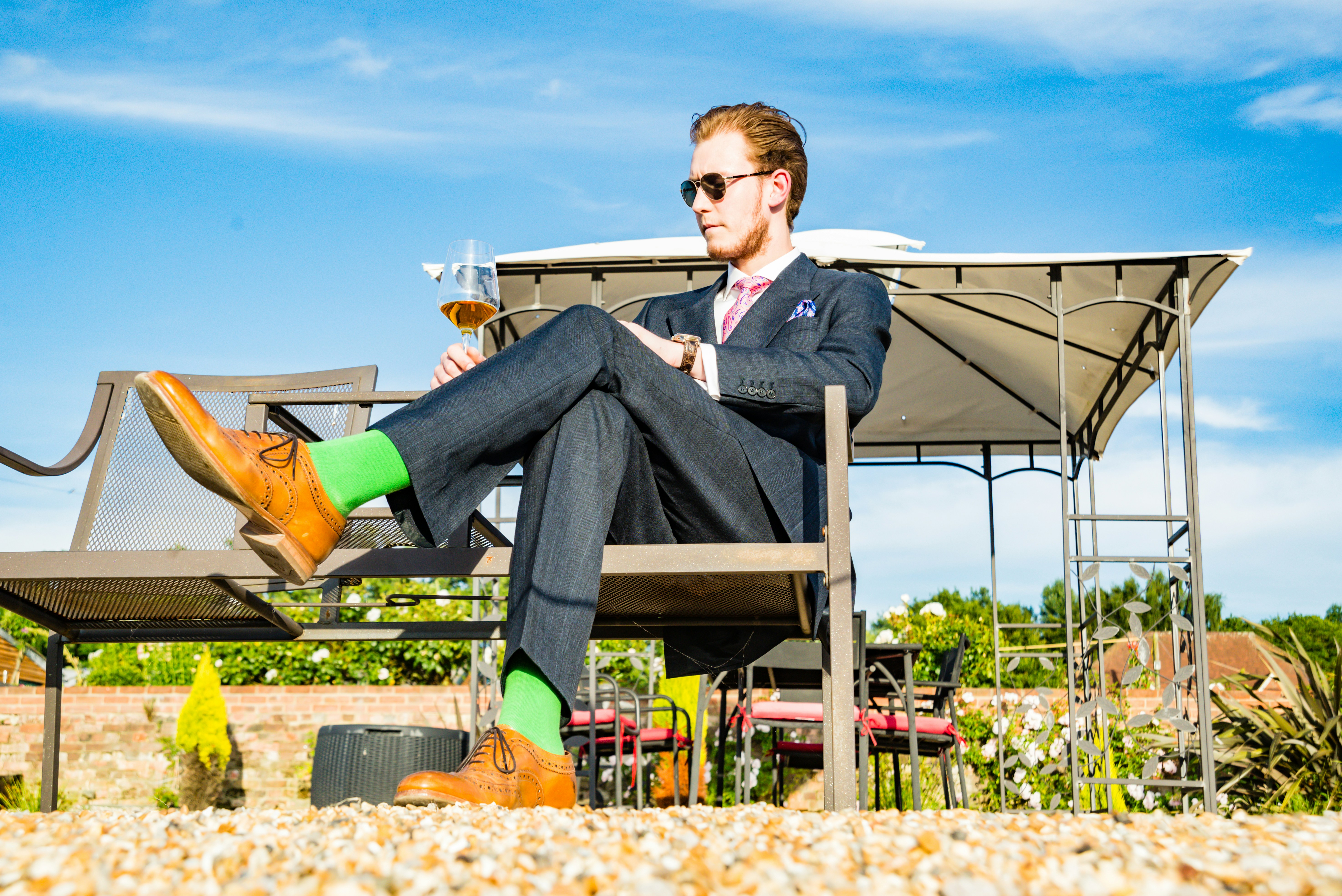 A man in sunglasses and a formal suit sitting on a bench outdoors with a glass of wine in his hand