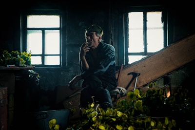 A reflective man is seated in a dimly lit room with two illuminated windows behind him, holding a cigarette close to his mouth. The room appears cluttered with various objects, including green leaves and a walking cane. The ambiance suggests an older, perhaps neglected setting.