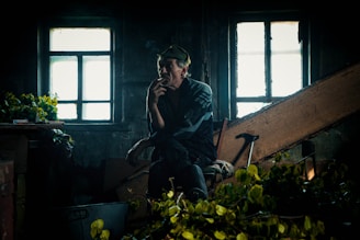 A reflective man is seated in a dimly lit room with two illuminated windows behind him, holding a cigarette close to his mouth. The room appears cluttered with various objects, including green leaves and a walking cane. The ambiance suggests an older, perhaps neglected setting.