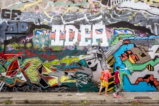 A vibrant urban graffiti wall with an eclectic mix of colors and shapes, featuring abstract designs and a large painting of a figure wearing a visor. Two people are walking along the sidewalk in front of the wall, one wearing a pink shirt and the other in yellow shorts with a red bag.