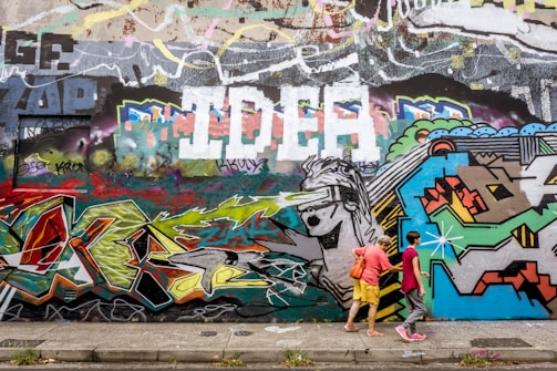 A vibrant urban graffiti wall with an eclectic mix of colors and shapes, featuring abstract designs and a large painting of a figure wearing a visor. Two people are walking along the sidewalk in front of the wall, one wearing a pink shirt and the other in yellow shorts with a red bag.