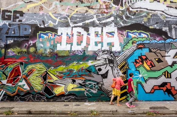A vibrant urban graffiti wall with an eclectic mix of colors and shapes, featuring abstract designs and a large painting of a figure wearing a visor. Two people are walking along the sidewalk in front of the wall, one wearing a pink shirt and the other in yellow shorts with a red bag.