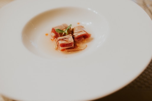 A minimalist plate features carefully arranged slices of tuna garnished with a delicate herb sprig and thin strands. The dish is presented on a large, elegant white plate, emphasizing the fine dining aesthetic.
