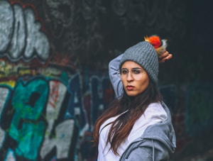 A person in a gray beanie with a colorful pom-pom strikes a confident and serious pose. The individual is wearing glasses and a casual gray hoodie over a white shirt. In the background, vibrant and colorful graffiti covers a dark wall, enhancing the street-like atmosphere.
