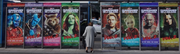 Large colorful posters of movie characters lined up at a bus stop advertising a film titled 'Guardians of the Galaxy Vol. 2'. A woman is seen walking in front of the posters.