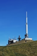 A transmission tower and several smaller communication structures are situated on a grassy hill under a clear blue sky. The large tower, with its long white antenna, dominates the scene, accompanied by smaller buildings with various antennas and satellite dishes.