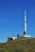 A transmission tower and several smaller communication structures are situated on a grassy hill under a clear blue sky. The large tower, with its long white antenna, dominates the scene, accompanied by smaller buildings with various antennas and satellite dishes.