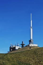 A transmission tower and several smaller communication structures are situated on a grassy hill under a clear blue sky. The large tower, with its long white antenna, dominates the scene, accompanied by smaller buildings with various antennas and satellite dishes.
