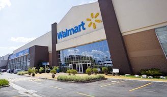 The image depicts the exterior of a large retail store with the Walmart logo prominently displayed. The building has a modern design with tall glass windows and is surrounded by a well-maintained parking lot and landscaped areas. Several parked cars are visible in front of the store.