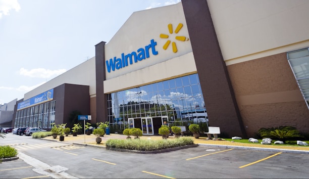 The image depicts the exterior of a large retail store with the Walmart logo prominently displayed. The building has a modern design with tall glass windows and is surrounded by a well-maintained parking lot and landscaped areas. Several parked cars are visible in front of the store.