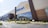 The image depicts the exterior of a large retail store with the Walmart logo prominently displayed. The building has a modern design with tall glass windows and is surrounded by a well-maintained parking lot and landscaped areas. Several parked cars are visible in front of the store.