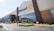 The image depicts the exterior of a large retail store with the Walmart logo prominently displayed. The building has a modern design with tall glass windows and is surrounded by a well-maintained parking lot and landscaped areas. Several parked cars are visible in front of the store.