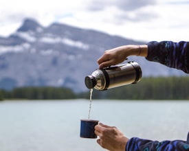 A person pours liquid from a silver thermos into a small cup, with a scenic backdrop of a large lake and mountains in the distance. The person is wearing a patterned sweater.