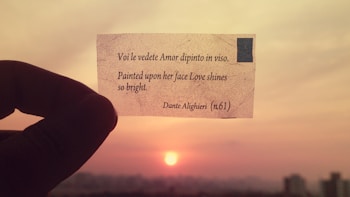 A hand holding a small piece of paper with a poetic line written on it against the backdrop of a sunset. The sky is a mix of orange and pink hues, and the sun is visible near the horizon.
