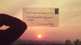 A hand holding a small piece of paper with a poetic line written on it against the backdrop of a sunset. The sky is a mix of orange and pink hues, and the sun is visible near the horizon.