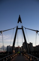 A bridge with a modern design stretches towards a cityscape of tall, residential buildings. The sky is clear, transitioning from a lighter shade near the horizon to a darker blue above. Several people are walking on the bridge, including one wearing a bright yellow-green jacket. The lighting is soft, suggesting either early morning or late evening.