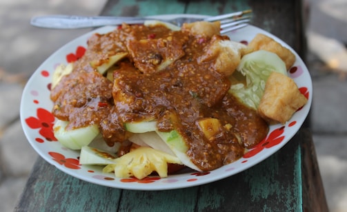 A colorful plate of traditional Indonesian salad known as gado-gado is topped with a rich peanut sauce. The dish includes fresh vegetables such as cucumber, and chunks of fried tofu are visible.