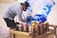 A person wearing a cap and mask is spray painting on a wall with a variety of colorful spray paint cans on a cart nearby. The background art features swirling patterns in blue and purple, suggesting creative expression.
