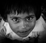 A close-up portrait of a young child with expressive eyes, looking directly at the camera. The child has short, dark hair and is wearing a collared shirt. The image is in black and white, highlighting the contrast and details of the child's facial features.
